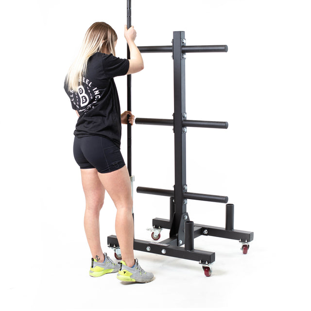 A woman with long blonde hair adjusts the Bells of Steel Bumper Plate Weight Tree and Bar Holder in black metal. She wears a black t-shirt, shorts, and light sneakers. The wheeled rack has horizontal bars, ideal for bar storage systems, against a white backdrop.