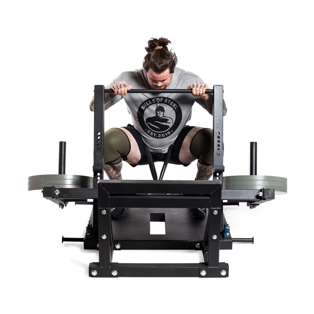 A person with long hair pulled back lifts weights on a Bells of Steel Belt Squat Machine to enhance lower body strength. Wearing a gray shirt with a circular logo and knee sleeves, they stand against a plain white background showcasing the space-efficient setup.