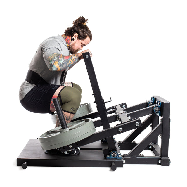 A tattooed, ponytailed person intensely focuses on a Bells of Steel Belt Squat Machine for lower body strength. Wearing a gray shirt, knee sleeve, and weightlifting belt, they exercise amidst large weights in their home gym.