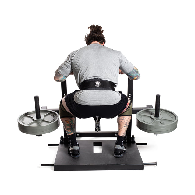 A tattooed individual enhances lower body strength using the Bells of Steel Belt Squat Machine. Wearing a gray shirt, black shorts, and a lifting belt in their home gym, they are viewed from behind against a crisp white background.