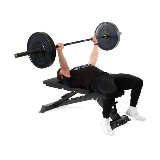 In a gym, someone in a black t-shirt and pants uses the Buzz-Saw Heavy-Duty Adjustable Bench by Bells of Steel to confidently perform a bench press with a barbell. The bench boasts an impressive 1,000lb max capacity.