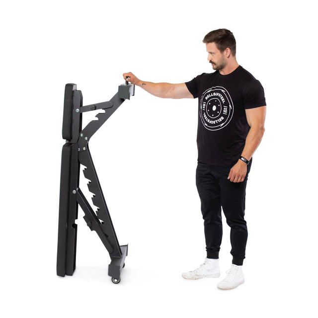 A person in a black t-shirt and pants stands next to the Buzz-Saw Heavy-Duty Adjustable Bench by Bells of Steel, highlighting its easy mobility with wheels and a 1000lb max capacity.