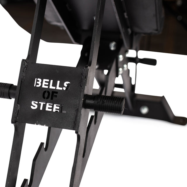 The image showcases the Buzz-Saw Heavy-Duty Adjustable Bench by Bells of Steel, highlighting its robust metal frame and bolts with a 1,000lb max capacity. An adjustable back pad adds versatility. The "BELLS OF STEEL" emblem is cut out on the bench, while the background remains out of focus.