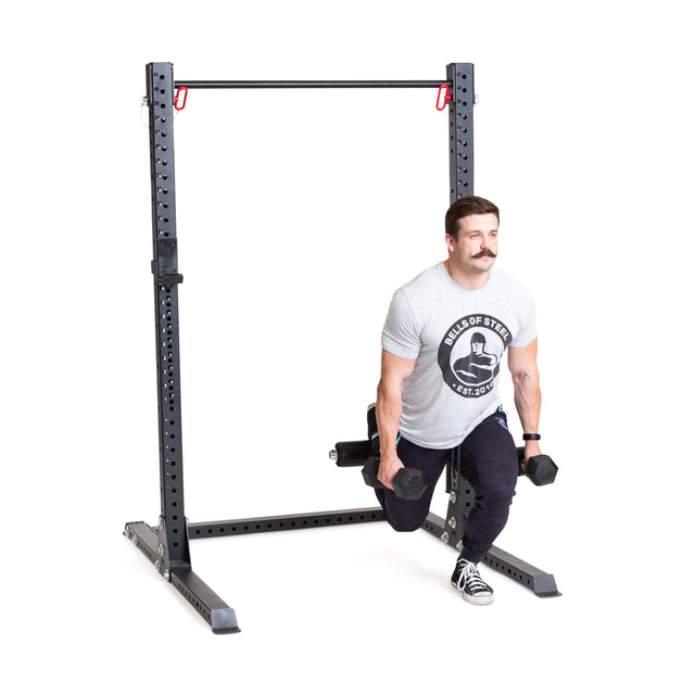 A man with a mustache and short hair performs a Bulgarian split squat using the Bells of Steel Split Squat Leg Roller Rack Attachment, while holding dumbbells, wearing a gray T-shirt and black pants, focused on perfecting his form.