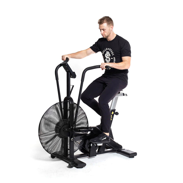 In a home gym, someone works out on a Bells of Steel Blitz Air Bike, donning black T-shirt and pants. The fan bike showcases a fan wheel and adjustable handles, all set against a plain white background.