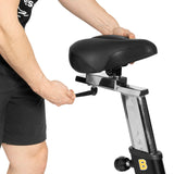 A person adjusts the height of the Blitz Air Bike seat by turning a knob on the metal post, getting ready for a full-body workout with the Bells of Steel bike in their home gym.