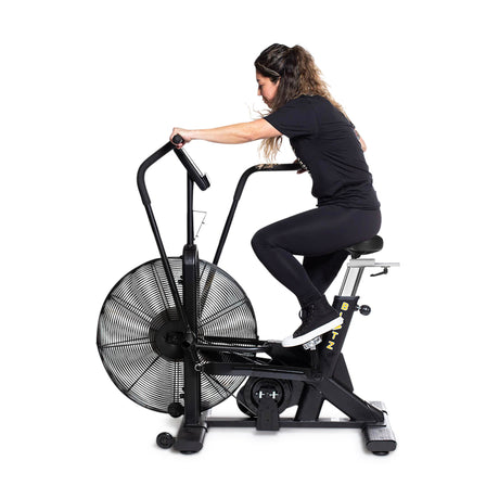 A woman in black athletic wear uses the Bells of Steel Blitz Air Bike, pedaling and pushing the handlebars for a full-body workout against a plain white background.