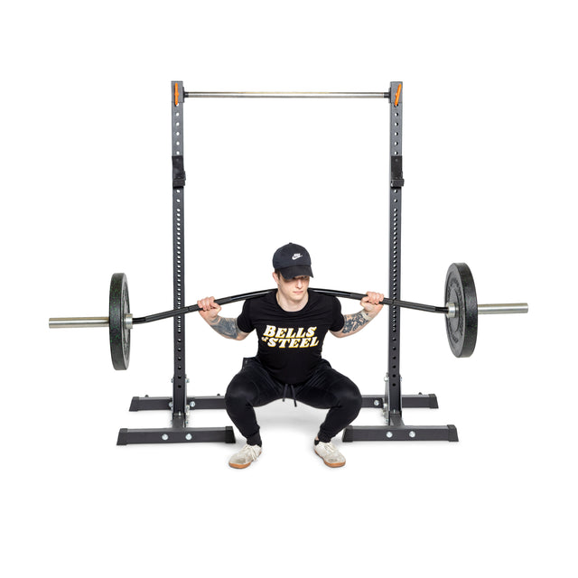 A person is squatting with a Bells of Steel Curved Squat Bar on their shoulders in a squat rack, wearing a black shirt with yellow text, black pants, a cap, and athletic shoes. The plain white background emphasizes their focus on muscular development.