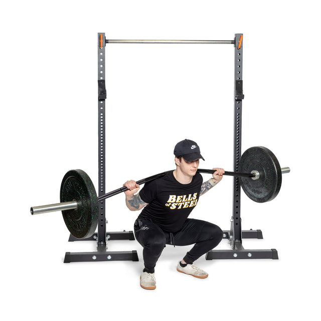 A person squats with a Bells of Steel Curved Squat Bar, enhancing muscle growth and reducing joint strain. Dressed in a black cap, shirt, and athletic shoes, they confidently face the rack with the barbell on their shoulders.