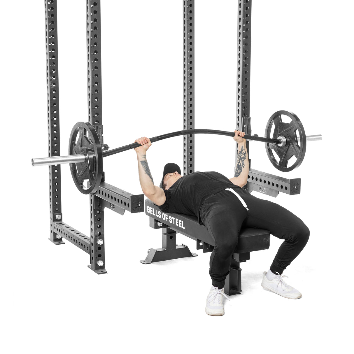 Clad in a cap and workout gear, a person bench presses on a Bells of Steel bench using their Buffalo Bar to lessen joint strain. Inside the power rack, they lift with determination, tattoos visible on their left arm, focusing intently on muscle development.