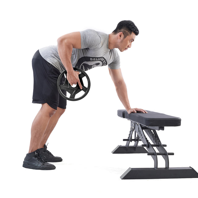 An athlete wearing sportswear performs a bent-over row with a Black Mighty Grip Olympic Weight Plate by Bells of Steel, stabilizing one hand on a bench against a white background.