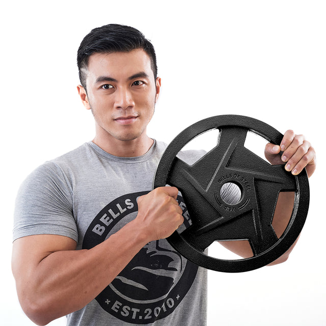 A man wearing a gray t-shirt with Bells of Steel's circular logo holds a Black Mighty Grip Olympic Weight Plate, highlighting its ergonomic design. The plain white background accentuates the precision grip handles for comfort and control.