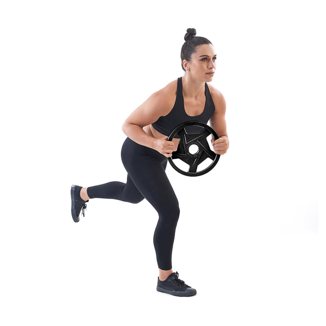 Against a white background, a person in a black workout outfit and sneakers performs a balance exercise on one leg, holding Bells of Steel's Black Mighty Grip Olympic Weight Plate with precision grip handles.