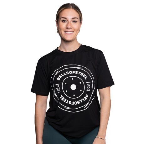 A smiling woman wears a black Bells of Steel Bamboo T-Shirt featuring a large white "BELLSOFSTEEL 25 KG" barbell plate graphic on the front, standing against a plain white background.