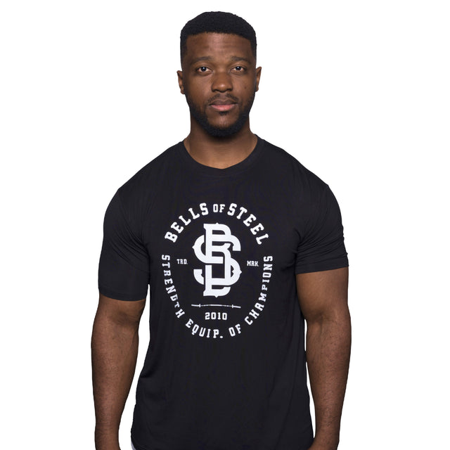 A person wearing the Bells of Steel Bamboo T-Shirt stands against a white background. The black shirt features the white "Bells of Steel" logo and "Strength Equip. of Champions" text. Made from sustainable resources, this buttery soft bamboo shirt offers optimal comfort and performance.
.