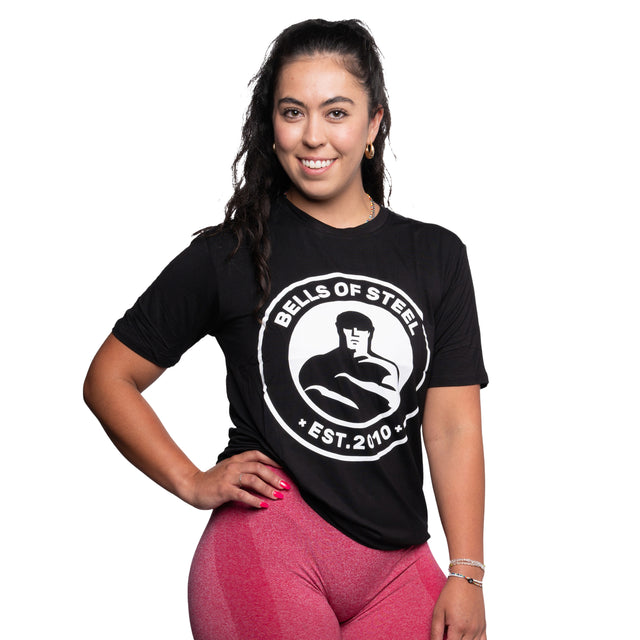 A woman in a Bells of Steel bamboo T-shirt and pink leggings smiles at the camera, her long dark hair contrasting the white background.