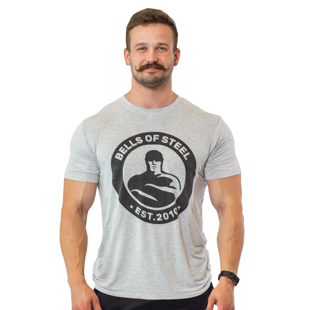 A man with a mustache smiles, wearing a gray Bells of Steel Bamboo T-Shirt featuring a muscular arm graphic and "EST. 2010." Its buttery soft bamboo blend ensures comfort and highlights sustainability. He has short hair against a plain white background.