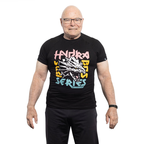 An older man with glasses and a smartwatch smiles against a white background, wearing the Bells of Steel Bamboo T-Shirt—a black sustainable workout shirt with a dragon graphic and colorful "Hydra Bass Series" text.
