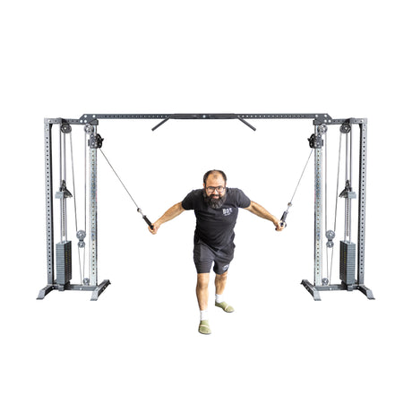 A man in a black shirt and shorts works out on the Bells of Steel Cable Crossover Machine, pulling the cables towards himself in a gym setting.