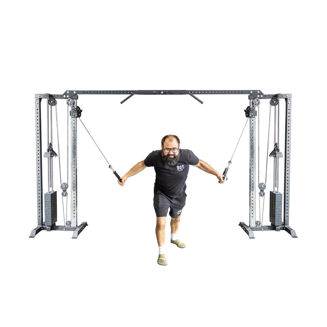 A man exercises on a Bells of Steel Cable Crossover Machine, dressed in a black T-shirt and shorts. He grips the adjustable handles, pulling the cables forward with both arms against a plain white backdrop.