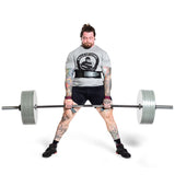 A muscular, tattooed man in a Bells of Steel shirt and shorts performs a heavy sumo deadlift using Machined Iron Olympic Weight Plate Sets by Bells of Steel against a plain white background.
