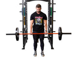 A man in a black graphic T-shirt and pants stands before a power rack, gripping a barbell with hands coated in Bells of Steel Mighty Grip Workout Chalk, ready to lift. The background is white and the gym equipment is visible behind him.