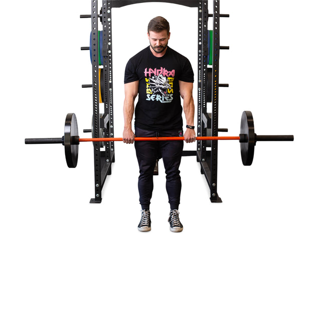 A man in a black graphic T-shirt and pants stands before a power rack, gripping a barbell with hands coated in Bells of Steel Mighty Grip Workout Chalk, ready to lift. The background is white and the gym equipment is visible behind him.