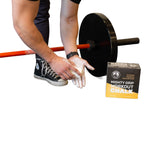 A person applies magnesium carbonate chalk to their hands near a barbell and a Bells of Steel Mighty Grip Workout Chalk box, all on a white background.