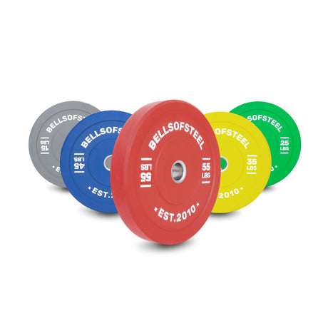 Five Bells of Steel Colored Bumper Plate Sets (gray, blue, red, yellow, green) with clear pound markings and stainless steel inserts are arranged diagonally on a white background.