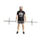 A person stands in a black T-shirt with a circular logo, shorts, and glasses, holding the Bells of Steel Hydra Seven Ring Bar. The plain white background highlights their powerlifting focus, complemented by black shoes.