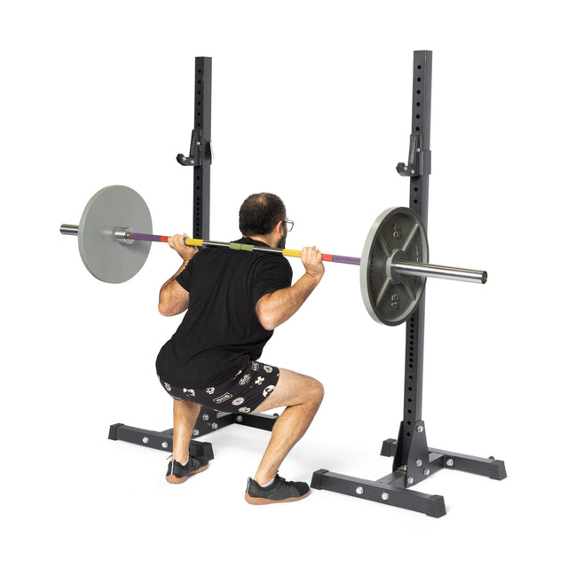 Wearing a black shirt, patterned shorts, and black shoes, an individual performs a squat with the Bells of Steel Hydra Seven Ring Bar. Positioned between a squat rack against a white background, they epitomize weightlifting prowess.