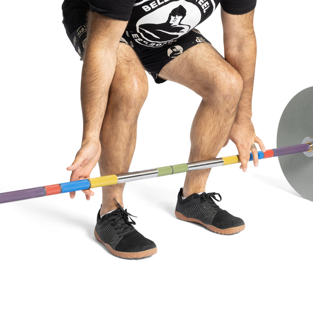 A person is lifting a barbell, wearing black athletic shoes and shorts, gripping the Bells of Steel Hydra Seven Ring Bar intently. The barbell, designed for powerlifting, features colorful markers and plates against a white background.