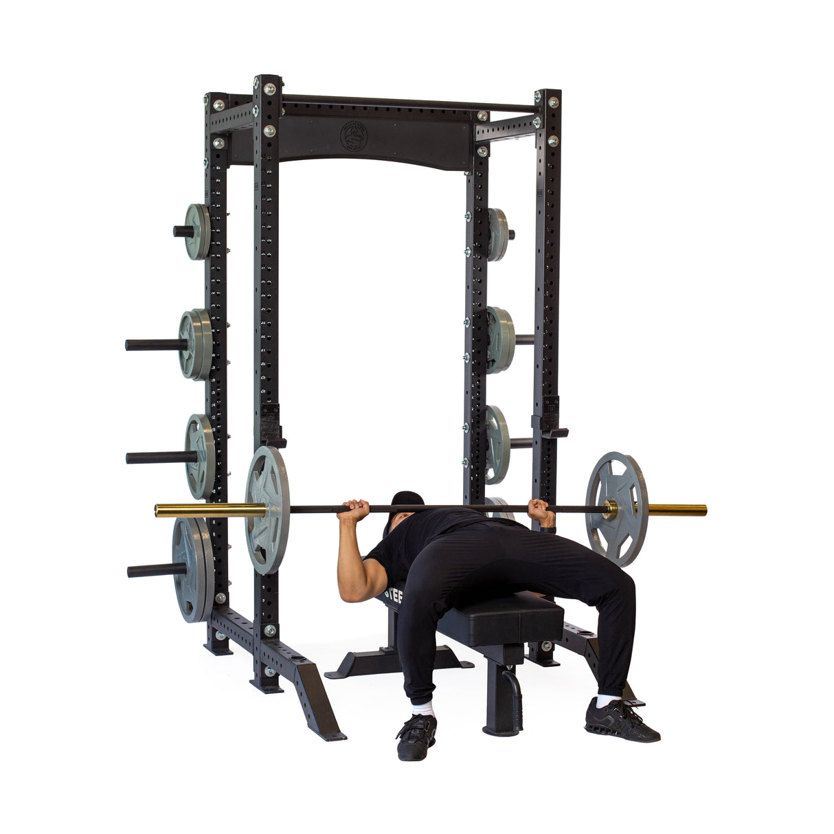 A person is performing a bench press under the Bells of Steel Hydra Collegiate Power Rack Builder (3" x 3", ⅝" Holes), lifting a barbell with weights. The rack, equipped with modular Hydra attachments, has extra weight plates stored on the sides. They're in black workout clothes and shoes.