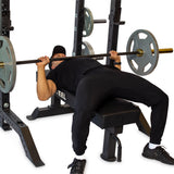 Dressed in black workout gear, an individual uses the Hydra Collegiate Power Rack Builder by Bells of Steel to perform a bench press. They grip the barbell loaded with weights on each side with enhanced stability from Hydra attachments.