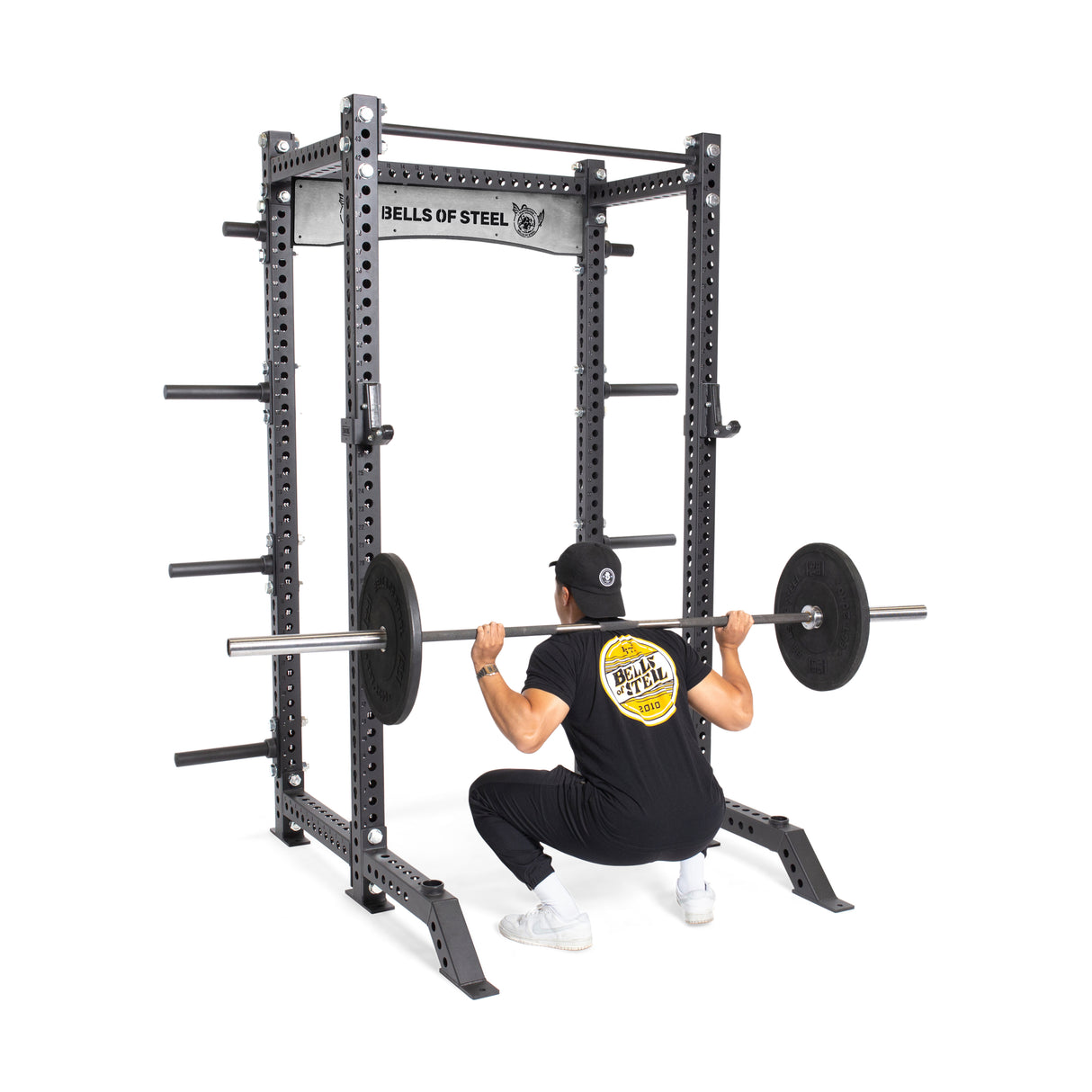 A person in a black T-shirt and cap is squatting with a barbell loaded with weights in the Bells of Steel Manticore Collegiate Power Rack Builder, which boasts a 1,000-pound capacity. The individual faces away, performing an impressive weightlifting exercise.