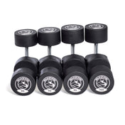 Five pairs of urethane dumbbells from Bells of Steel are arranged in a row, with weights labeled 85, 90, 95, and 100. Each boasts a logo on the end cap, highlighting their commercial-grade quality.