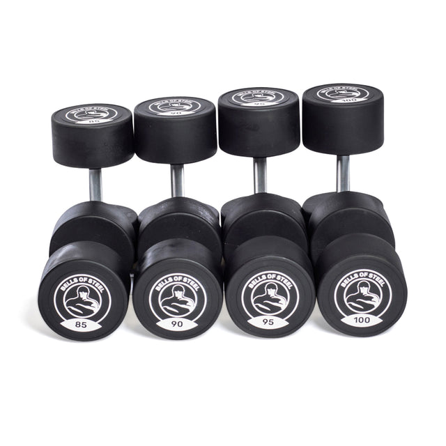Five pairs of Bells of Steel Urethane Dumbbell Sets, labeled 85–100 lbs, are arranged in a row with circular logos on the ends—demonstrating commercial-grade quality for serious lifters.