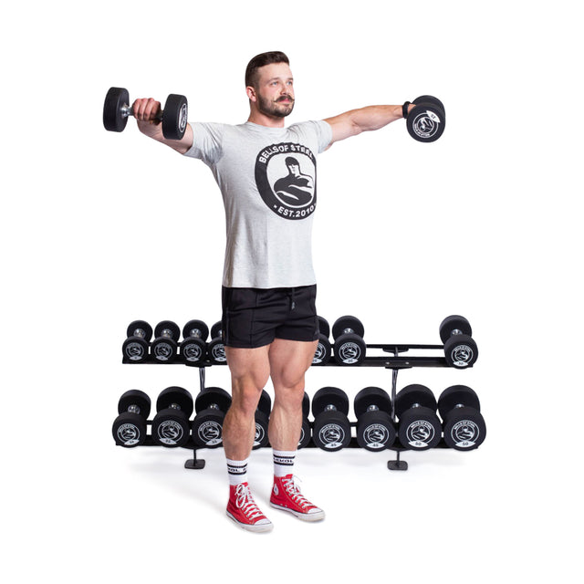 A man in a gray T-shirt and black shorts, wearing red sneakers and striped socks, holds Bells of Steel Urethane Dumbbells with arms extended. Behind him is a rack of commercial-grade equipment against a white background.