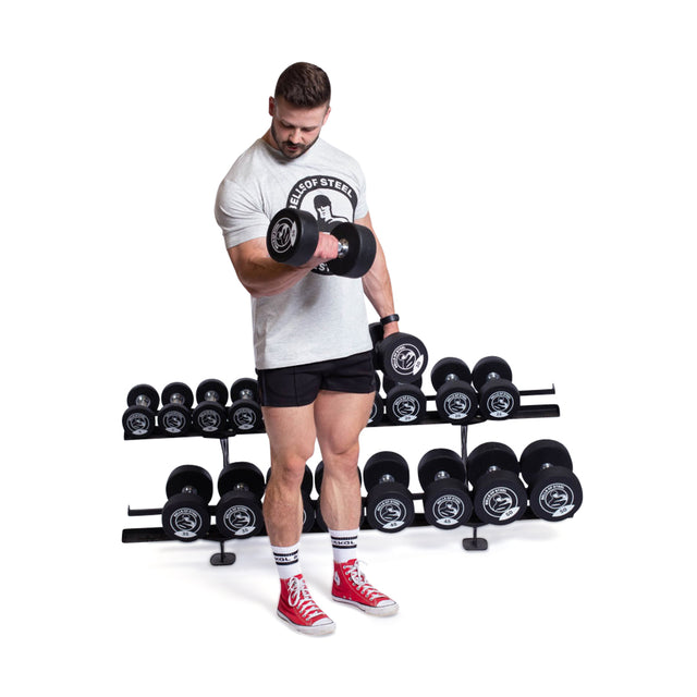 Clad in a light gray t-shirt, black shorts, and red high-top sneakers, an individual showcases the commercial-grade stainless steel head of a Bells of Steel dumbbell. Behind them is a rack of Urethane Dumbbells on the white floor.