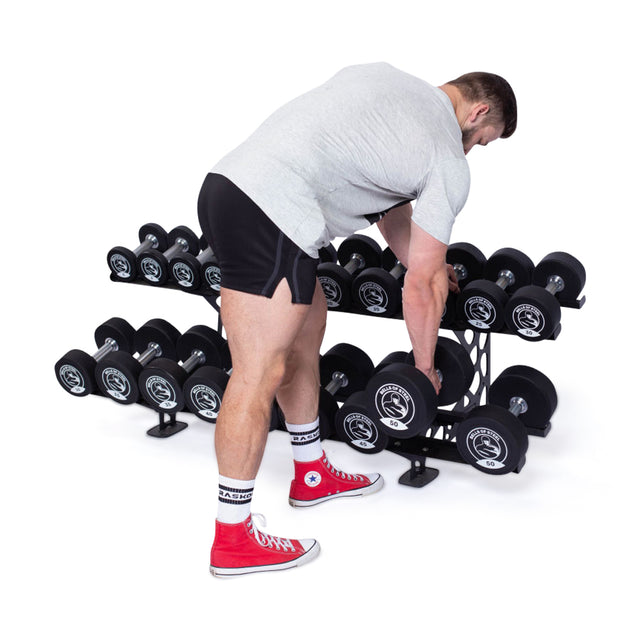 A person in a gray T-shirt, black shorts, and red sneakers is picking up a Bells of Steel Urethane Dumbbell from a commercial-grade rack displaying multiple rows of black dumbbells against a white background.