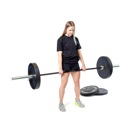 A woman in a black t-shirt and shorts lifts a barbell fitted with Bells of Steel Dead Bounce Conflict Bumper Plate Sets, made of virgin rubber, while more plates are stacked behind her on a white background.