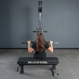 A person with long brown hair sits on a Bells of Steel bench, boosting grip strength while using the Bells of Steel Fat Bar - Olympic Curl Bar Cable Attachment with a lat pulldown, facing a gray wall and gym equipment in the workout room.
