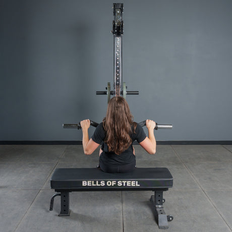 A person with long brown hair sits on a Bells of Steel bench, boosting grip strength while using the Bells of Steel Fat Bar - Olympic Curl Bar Cable Attachment with a lat pulldown, facing a gray wall and gym equipment in the workout room.