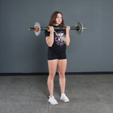 A woman in a black t-shirt and shorts stands indoors, holding the Bells of Steel Fat Bar - Olympic Curl Bar Cable Attachment at shoulder height to enhance grip strength. The background features a plain gray wall and tiled floor.