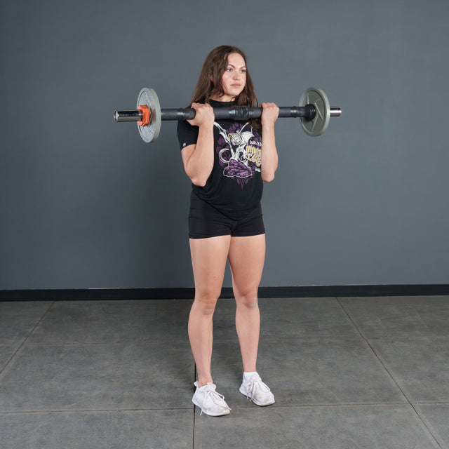 A woman in a black t-shirt and shorts stands indoors, holding the Bells of Steel Fat Bar - Olympic Curl Bar Cable Attachment at shoulder height to enhance grip strength. The background features a plain gray wall and tiled floor.