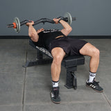 A person lies on a bench press bench, gripping the Bells of Steel Fat Bar - Olympic Curl Bar Cable Attachment above their chest. Wearing black workout gear and white socks, they're ready to test their grip strength.