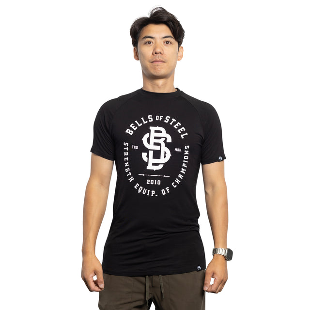 A person poses against a white background in a buttery soft black bamboo blend t-shirt, featuring "Bells of Steel" and "Strength Equip. of Champions," along with "2010" and a logo. This comfortable Bamboo T-Shirt is paired with khaki pants for a casual yet stylish look.