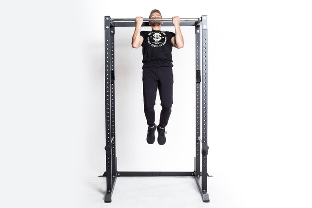 A man in black athletic wear performs a pull-up on the Bells of Steel Residential Power Rack (2.3" x 2.3", ⅝" holes), featuring metal j-cups, against a plain white background, highlighting home gym strength and versatility.