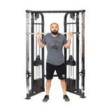 A man stands before a cable machine, gripping the Bells of Steel Double Hook Straight Bar Cable Attachment across his shoulders as he prepares for exercises. He wears a gray shirt, black shorts, and sneakers.