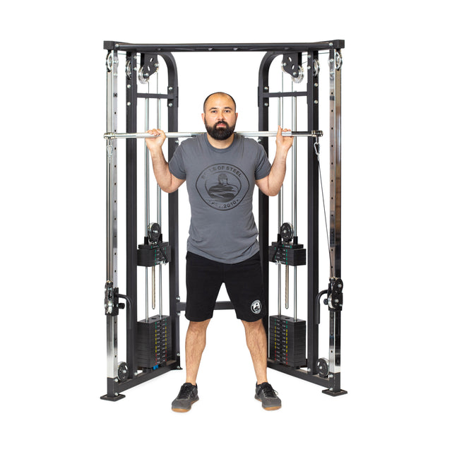 A man stands before a cable machine, gripping the Bells of Steel Double Hook Straight Bar Cable Attachment across his shoulders as he prepares for exercises. He wears a gray shirt, black shorts, and sneakers.